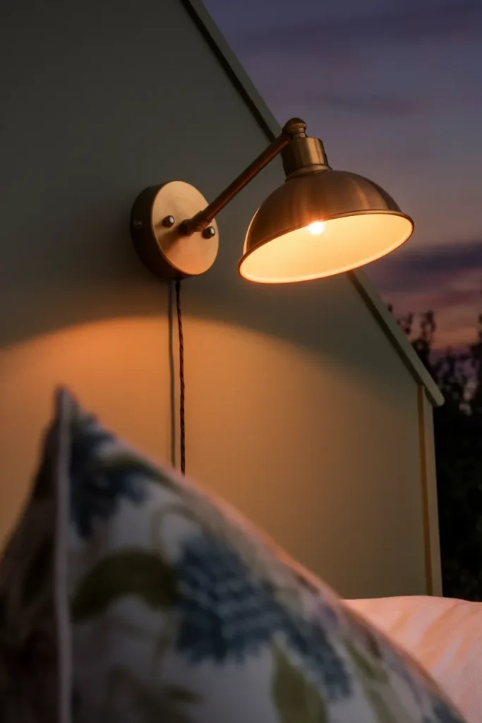 Plug-in brass swing-arm sconce glowing warm 2700K beside a cottagecore bed