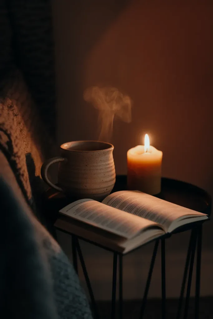 Steaming mug, lit beeswax candle, and face-down paperback on a side table in warm candlelight.