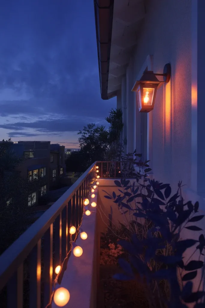 Balcony garden at blue hour lit by warm string lights and a plug-in wall lantern.
