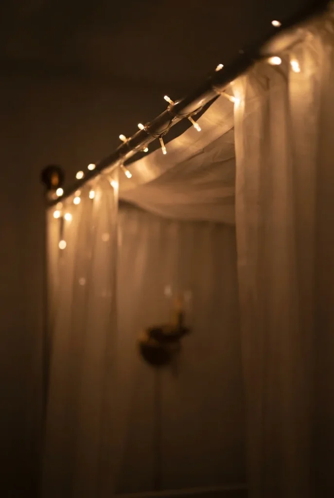 Warm string lights along a canopy rail glowing through sheer ivory bed curtains at night.