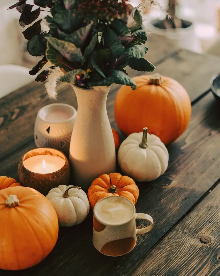 Cozy fall tabletop on dark wood with orange and white pumpkins, a lit candle in a coconut shell, a cream vase of ornamental kale and greenery, and a speckled ceramic mug of latte.