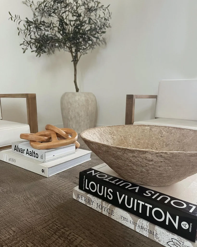 Dark wood coffee table styled with stacked design books, a wooden chain sculpture, and a rustic textured bowl, with an olive tree behind.