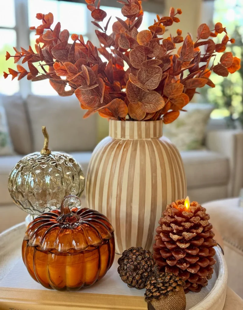 Fall coffee table vignette with a striped ceramic vase of rust-colored faux eucalyptus, an amber glass pumpkin jar, a mercury glass pumpkin, a lit pinecone-shaped candle, and decorative acorns on a white tray.