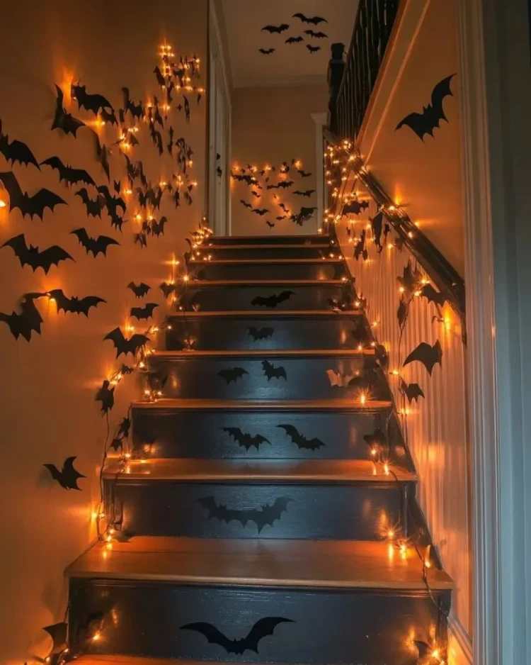Halloween-decorated staircase with black bat wall decals covering the surrounding walls and stair risers, lit by warm orange string lights along the banister and steps.