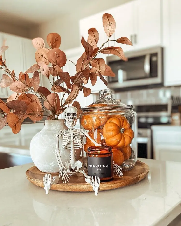Halloween kitchen counter vignette on a wooden tray with a decorative skeleton figure, a white vase of rust-colored faux eucalyptus, a glass jar filled with mini pumpkins, and a Cinnamon Rolls candle.