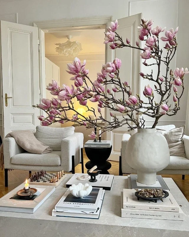 Pink magnolia branches in a white sculptural vase on a marble coffee table with stacked books, a chess set, and a candle.