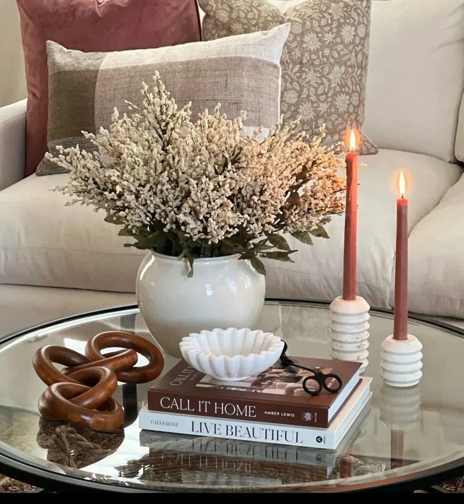 Round glass coffee table styled with dried cream florals in a white vase, lit terracotta taper candles, stacked design books, and a wooden chain sculpture.