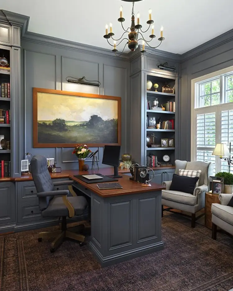 Traditional home office with slate blue paneled walls and built-in bookshelves, a wood-topped desk with a leather chair and computer monitor, a framed landscape painting, a chandelier, wingback armchairs, and a patterned area rug.