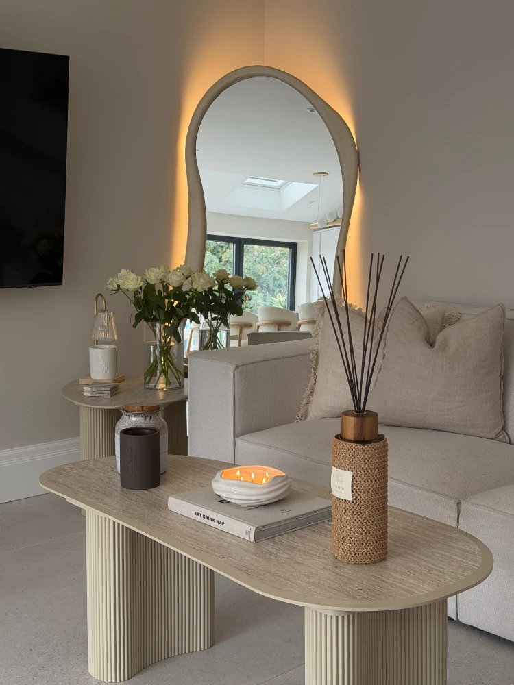 Travertine coffee table with a lit candle, reed diffuser, and book, beneath a backlit wavy mirror, with white roses and a beige sofa nearby.
