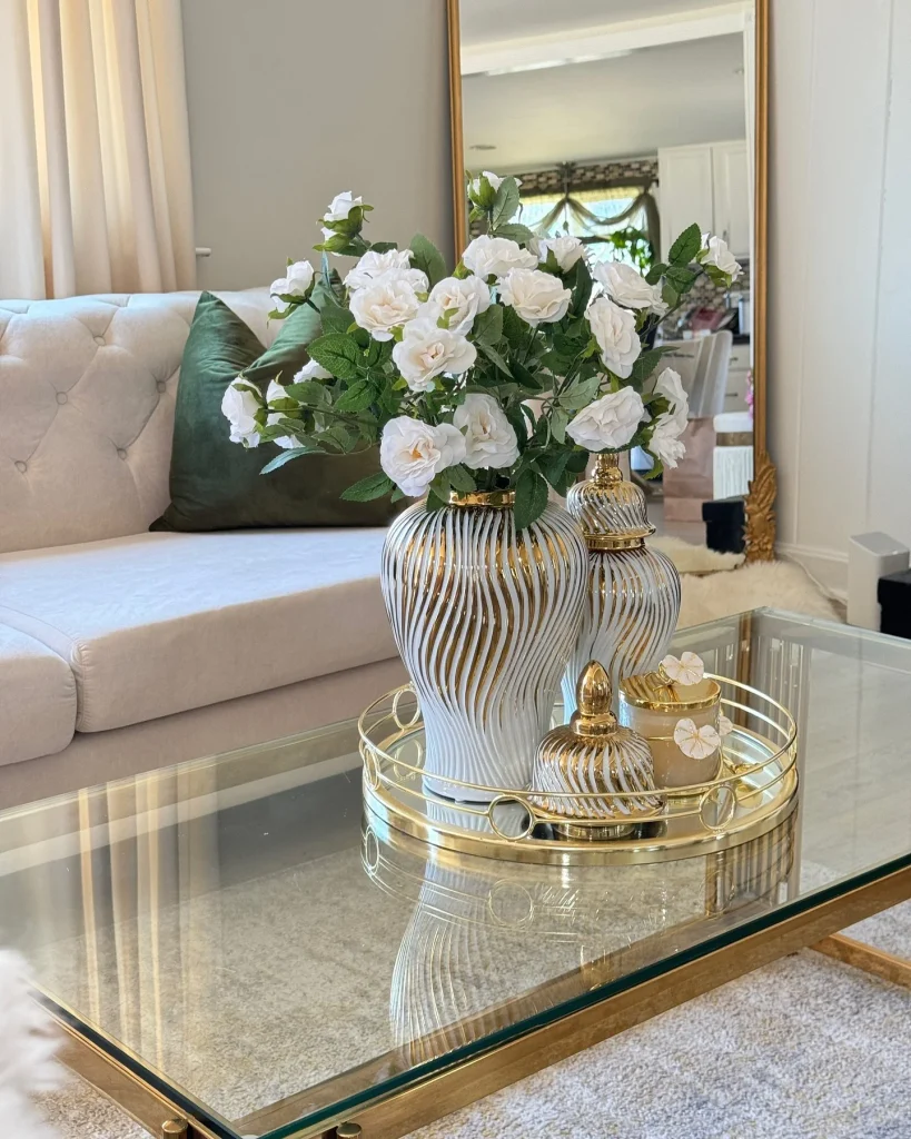 White and gold swirl ginger jars with white roses on a gold mirrored tray atop a glass coffee table, with a cream tufted sofa behind.