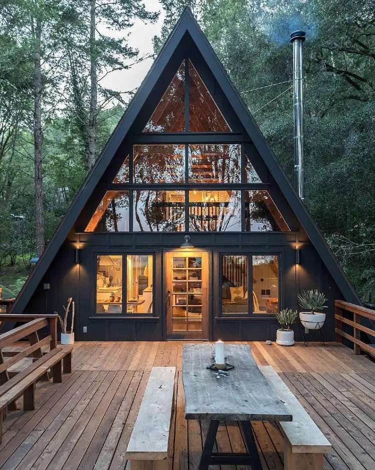 Black A-frame cabin with a triangular glass facade glowing warmly at dusk, a wood-plank deck with a picnic table and benches, potted plants, and a smoking chimney, surrounded by dense forest.