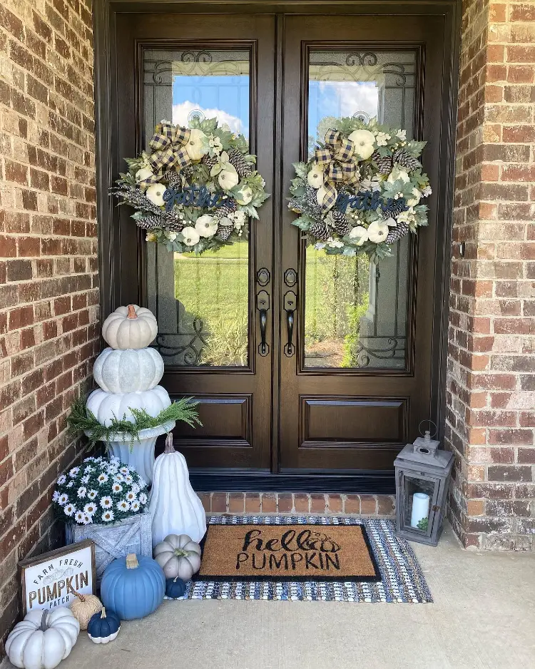 Wood-and-glass double front doors flanked by brick, each hung with a "gather" wreath of white pumpkins, plaid ribbon, and pinecones, with a stacked pumpkin topiary, blue and white pumpkins, a "Farm Fresh Pumpkin Patch" sign, a lantern, and a "Hello Pumpkin" doormat.