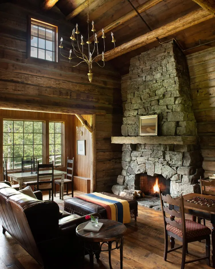 Wood cabin living room with a tall floor-to-ceiling stone fireplace lit with a fire, a brass candelabra chandelier, a dark leather sofa with a striped blanket, a wooden dining table by a window, and antique wooden chairs.