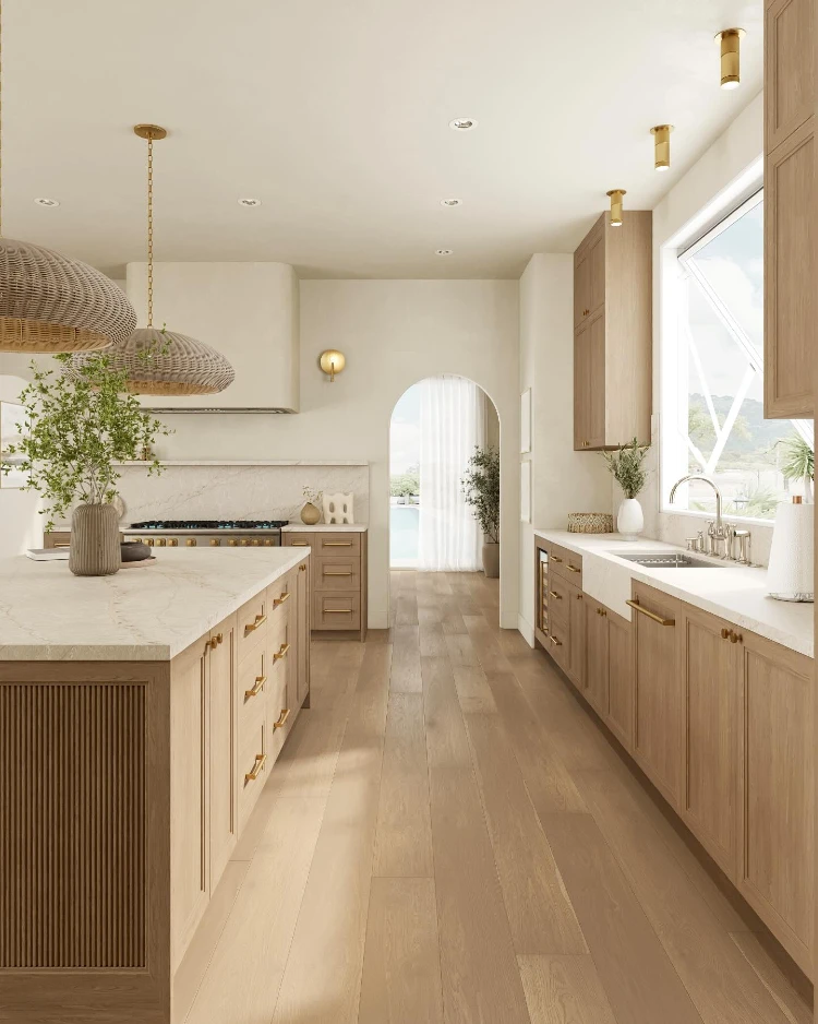 Bright coastal-modern kitchen with light oak cabinetry, a large island with fluted paneling, woven pendant lights, brass hardware, marble countertops, and an arched doorway leading to a sunlit hallway with a pool view.