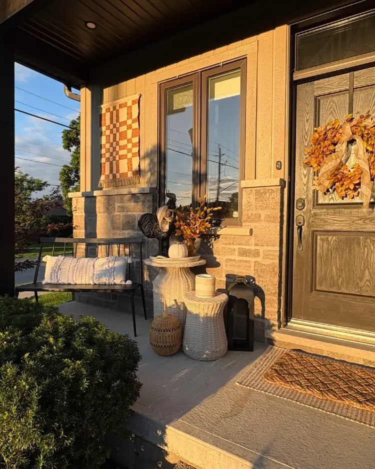 Sunlit front porch at golden hour with a wood-look door decorated with a burlap-tied fall wreath, a black metal bench with a pillow, woven side tables, a rooster figurine and pumpkins, a hanging patchwork quilt, and a jute doormat.