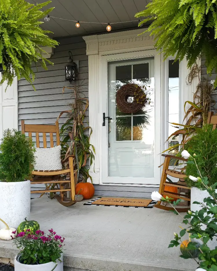 Grey farmhouse front porch with a white door decorated with a natural grapevine wreath, twin wooden rocking chairs flanking the entrance, hanging Boston ferns, cornstalks, pumpkins, magenta mums, and string lights overhead.