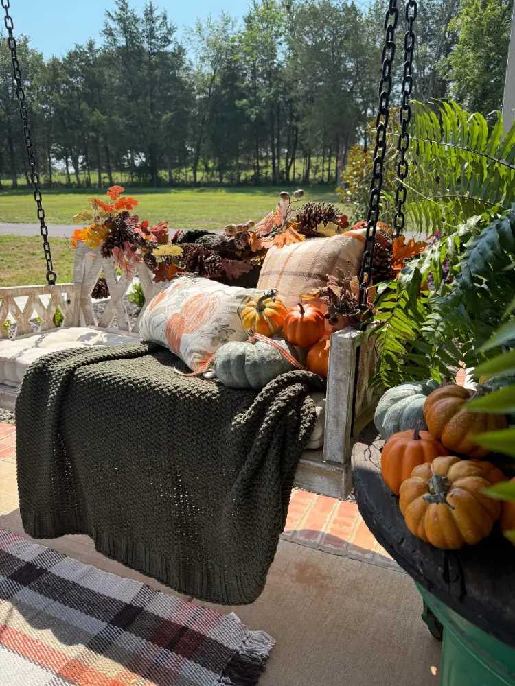 White wooden hanging porch swing draped with an olive knit throw, plaid and pumpkin-print pillows, fall foliage and pinecones, mini pumpkins and gourds, and a fern, overlooking a grassy field lined with trees.