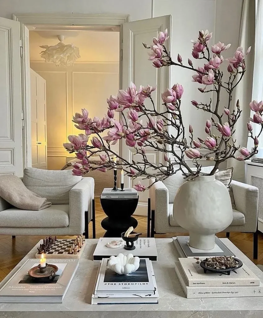 Pink magnolia branches in a white sculptural vase on a marble coffee table styled with stacked books, a chess set, and a lit candle, with cream armchairs and white paneled doors behind.