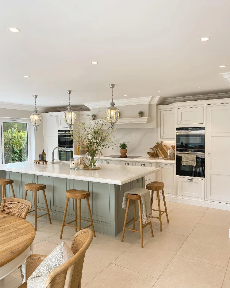 Spacious kitchen with a sage green island topped with white marble, wooden bar stools, glass pendant lights, white shaker cabinetry, built-in double ovens, and a round wood dining table with woven chairs in the foreground. Modern Farmhouse Kitchens