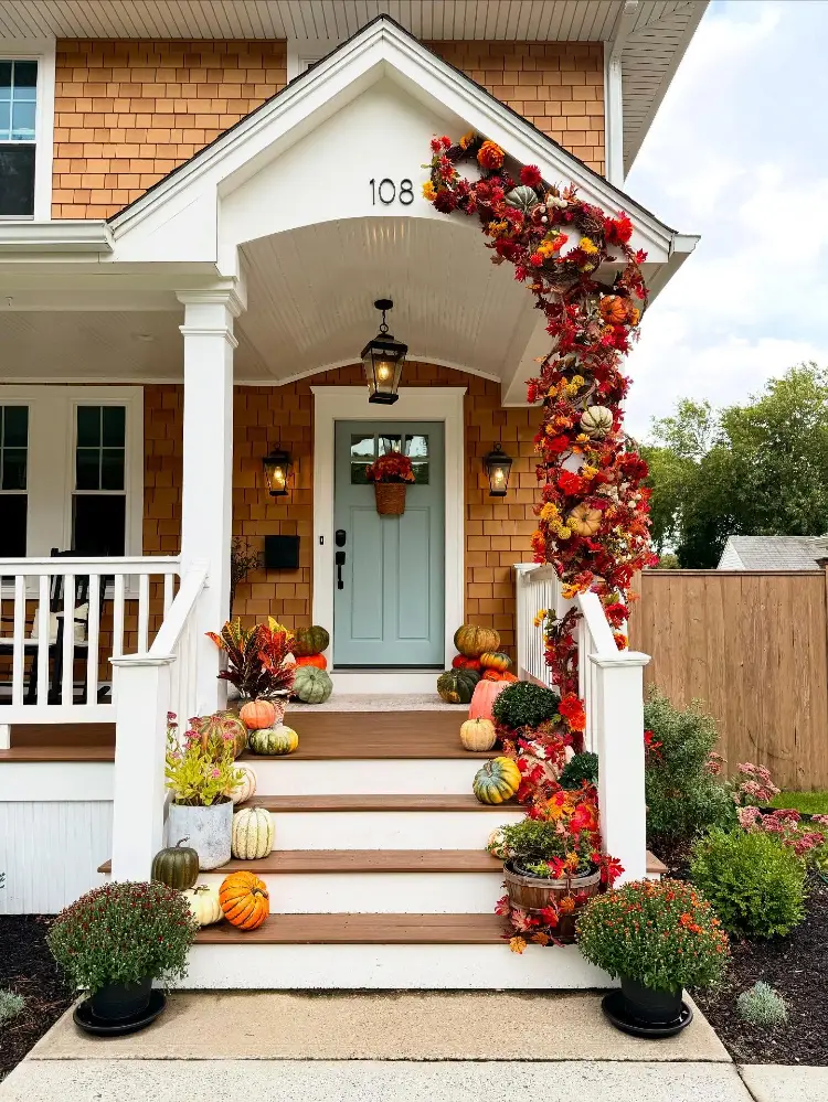 Shingle-style house entrance with a pale blue front door, white porch columns wrapped in a vibrant red and orange floral garland with pumpkins woven in, wooden steps lined with mixed pumpkins and mums, and lantern sconces.