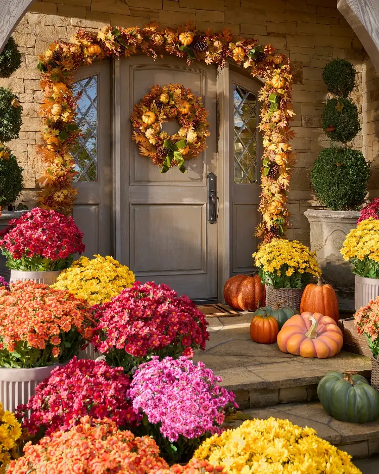 Stone house entrance with a taupe double door framed by a lush pumpkin and foliage garland archway and matching wreath, surrounded by an abundance of colorful mums in pink, red, orange, and yellow, and mixed heirloom pumpkins on stone steps.