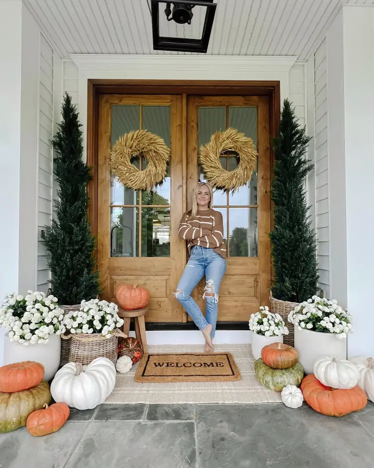 A woman in a striped sweater and ripped jeans leans against a wood double door decorated with woven wheat wreaths, surrounded by potted white mums, faux topiary trees, mixed pumpkins, and a "Welcome" doormat.