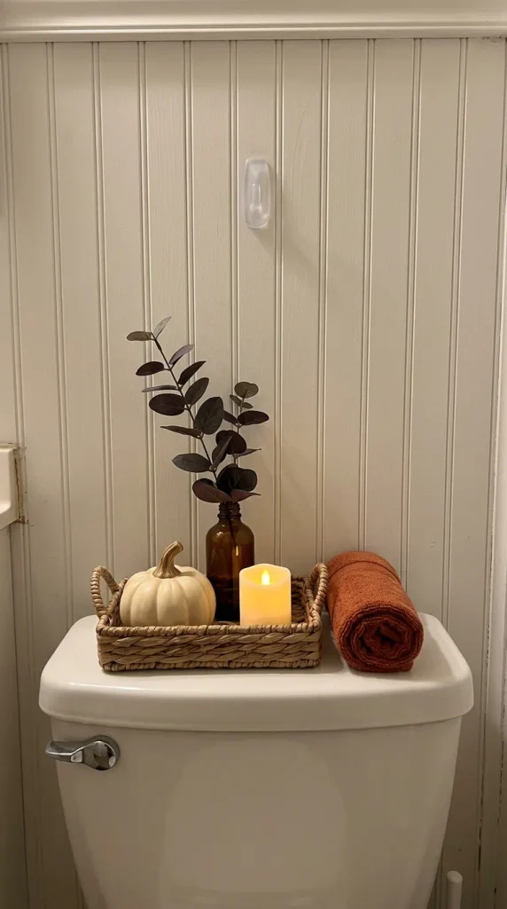 Small bathroom fall decor on a toilet tank tray with a ceramic pumpkin, amber bottle, and LED candle at night