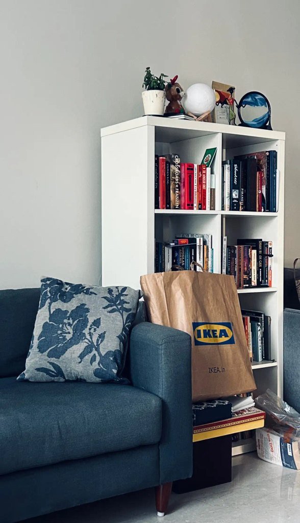 Teal-blue sofa with a floral pillow beside a white cube bookshelf and an IKEA shopping bag