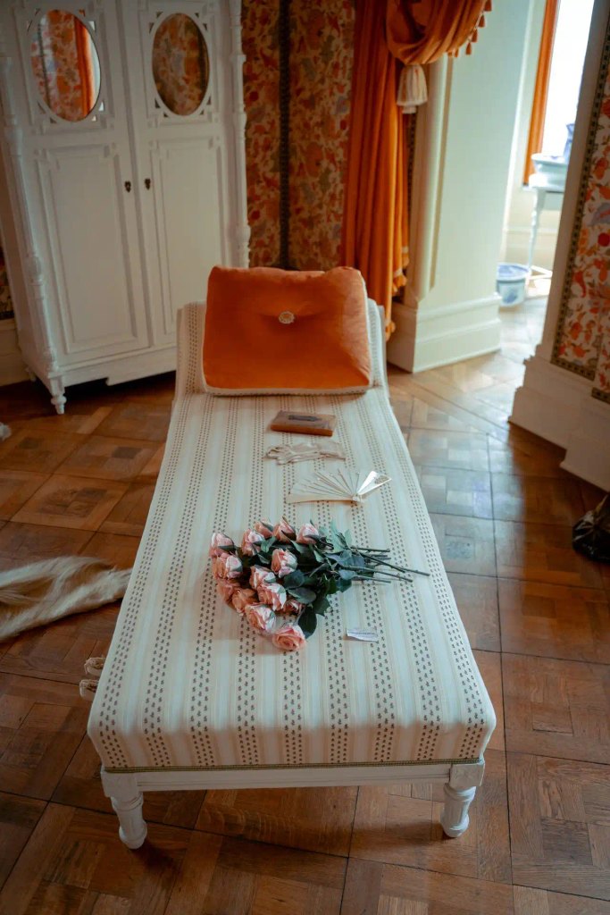 An antique chaise lounge with striped cream upholstery, an orange velvet bolster pillow